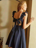 jeune femme portant une robe courte dos nu bleue evangeline avec des noeuds, posant près d'une fenêtre ensoleillée