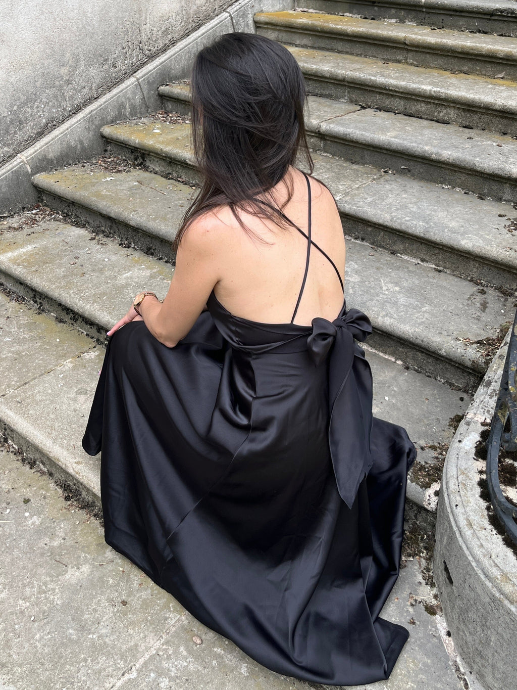 Femme assise portant une élégante robe satin mi-longue noire, dos nu, Tessa, sur des marches en pierre pour une allure chic.