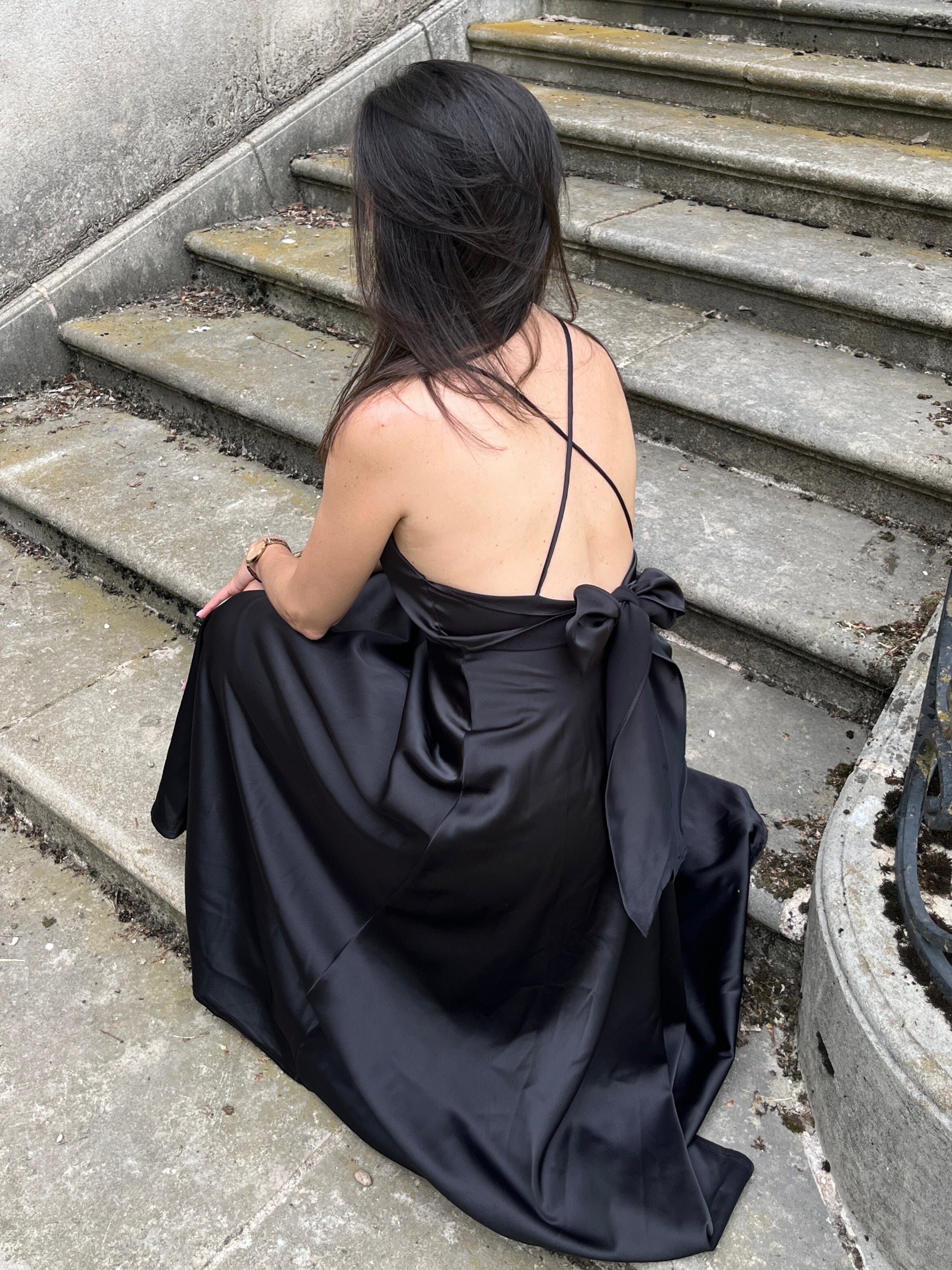 Femme assise portant une élégante robe satin mi-longue noire, dos nu, Tessa, sur des marches en pierre pour une allure chic.